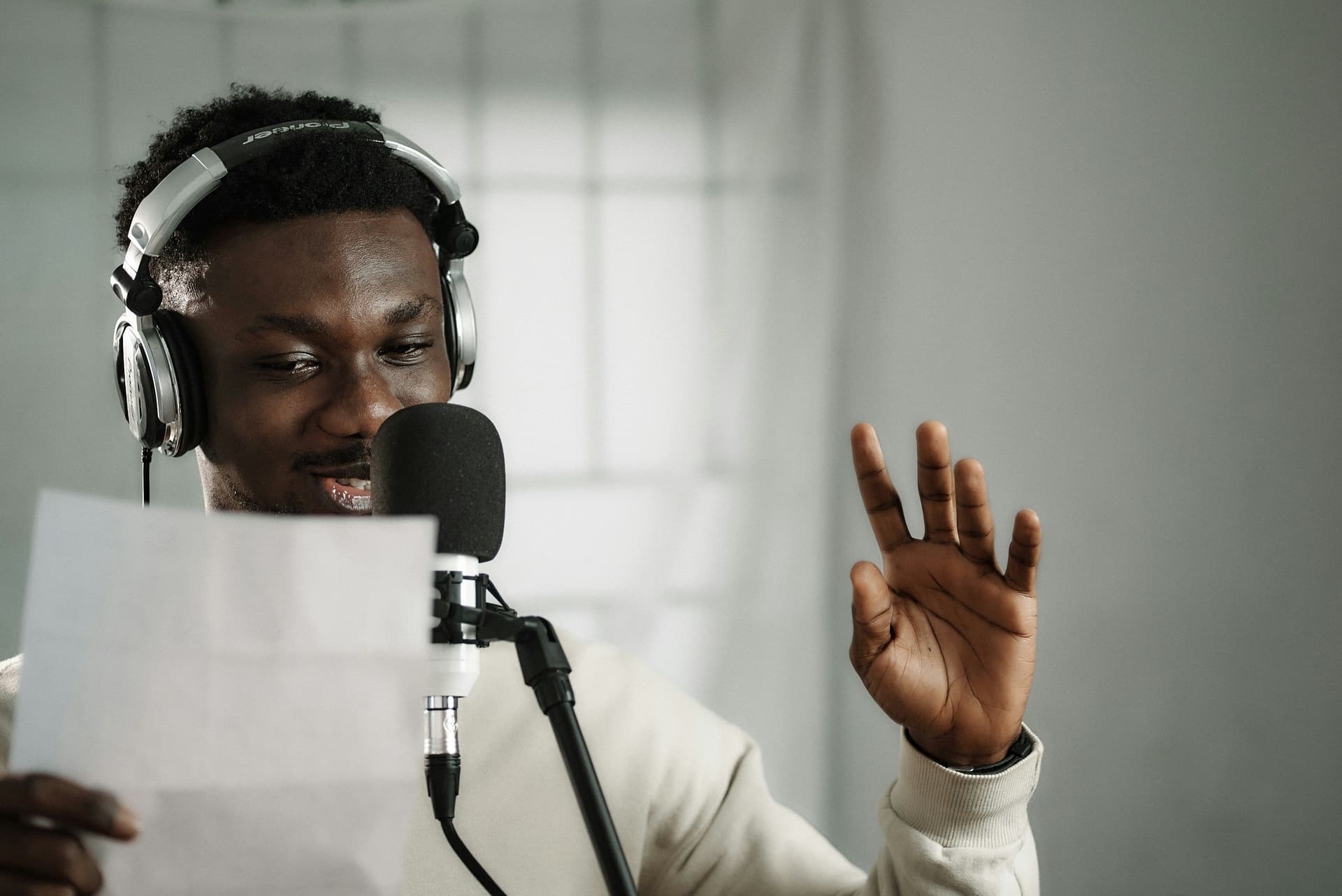 Person with headphones in front of a mic gesturing with hand while holding a paper.
